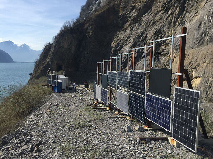 Solarpanels auf Kiesplatz an steilem Hang, links See und Berge