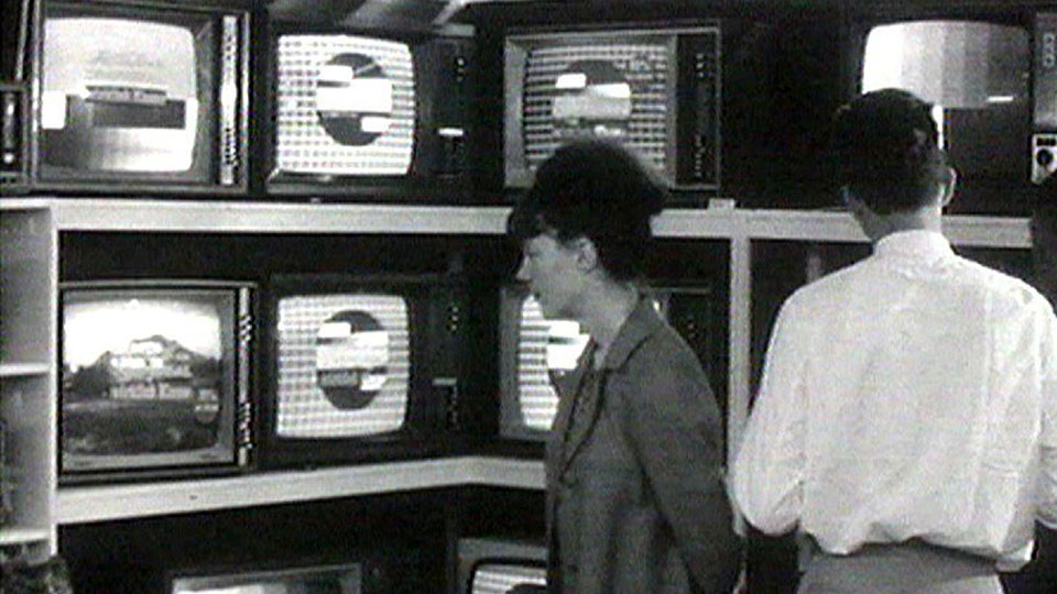 Ein Mann und eine Frau betrachten ausgestellte TV-Geräte (Schwarzweissfoto)