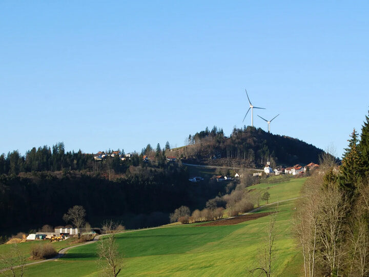 Bewaldeter Hügel, vereinzelte Häuser oder Höfe, im Hintergrund zwei Windkraftanlagen