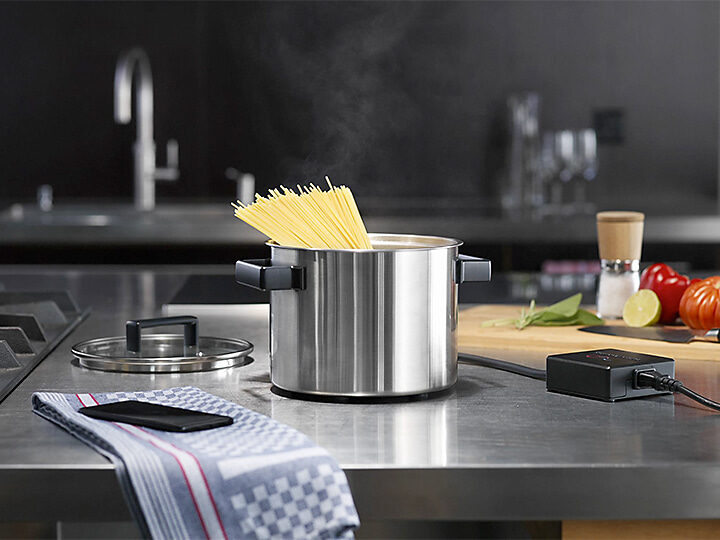 Topf mit Pasta auf Chromstahl-Küchentisch, rechts davon das Netzteil des Topfs, links auf einem Küchentuch liegt ein Smartphone
