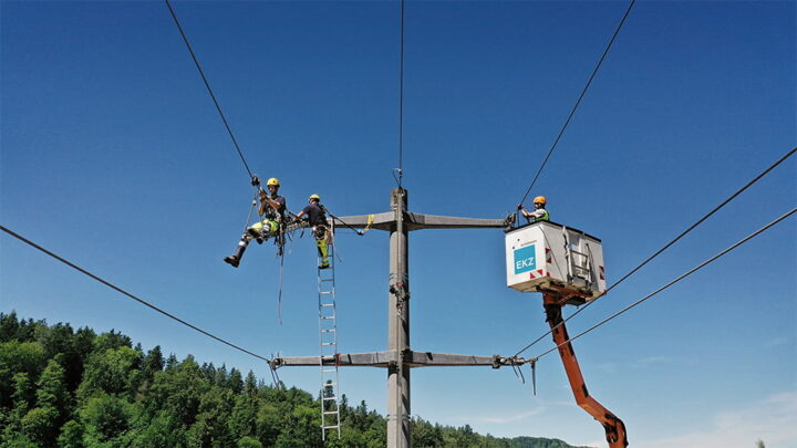 Drei Monteure arbeiten an einem Strommast, davon einer auf einer Leiter, einer im Korb einer Hebebühne, der Dritte nutzt die Leitung als Seilbahn