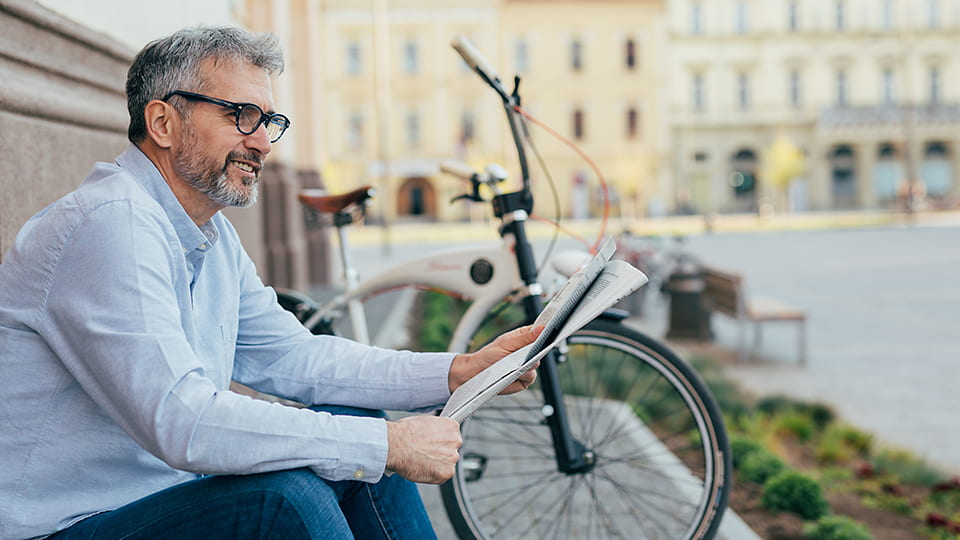 Ein Mann sitzt im Schatten und liest Zeitung, im Hintergrund ein Fahrrad und Bürogebäude