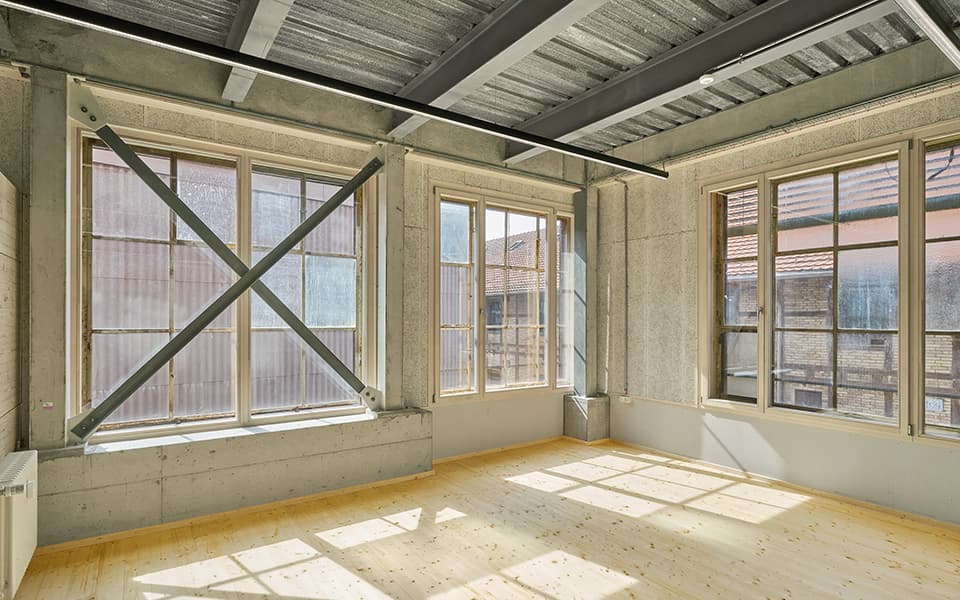 Ein Raum mit hellem Holzboden, Sichtbeton- und Lehmputzwänden, unterschiedlich aufgeteilten Fenstern sowie Stahlträgerdecke.