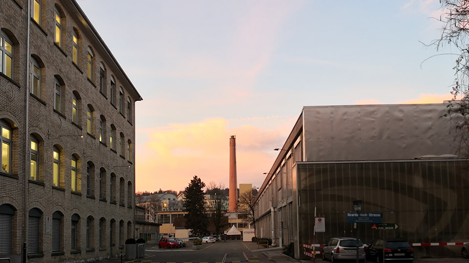 Fabrikgelände mit Altbau, Hochkamin und moderner Halle in heller Morgenstimmung – im Altbau brennt Licht